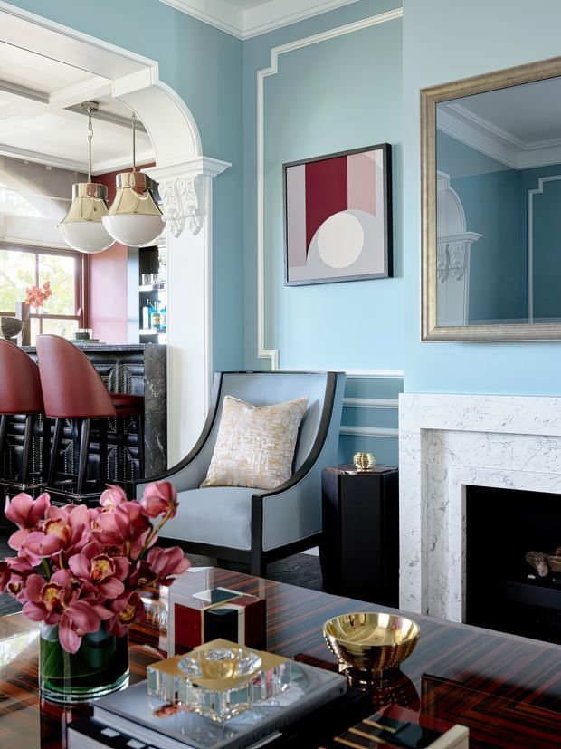 A refined, traditionally detailed living room with pale blue walls, ornate white decorative mouldings, a marble fireplace, a black-trimmed upholstered armchair, and a glossy dark coffee table. An arched opening leads to a bar or kitchen area with red upholstered stools and pendant lights. The room includes a large framed mirror, geometric artwork, and floral arrangements, creating a polished, colourful interior.