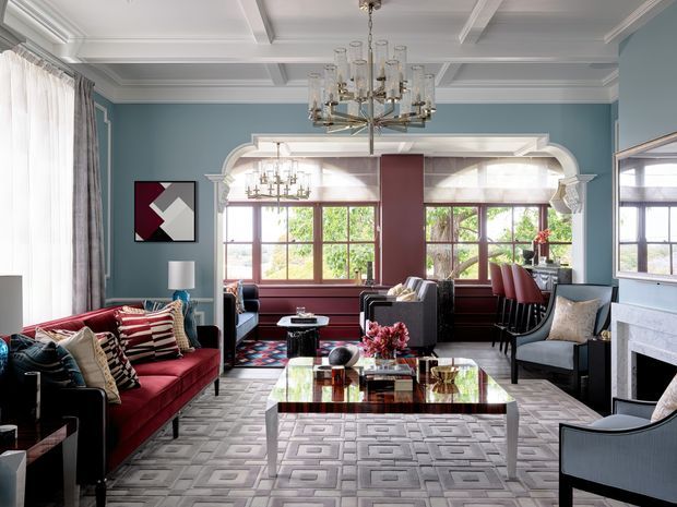 A refined living room with blue walls, elaborate white coffered ceiling and moulded archway, large timber-framed windows, a red velvet sofa, patterned rug, glass coffee table, sculptural chandelier, and a fireplace at right. The room combines classical architectural detailing with bold, tailored furnishings and a view into an adjoining dining or sitting area.