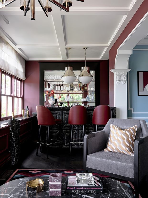A richly styled home bar and lounge corner with a black marble bar, three burgundy upholstered stools, mirrored glass shelving, decorative pendant lights, dark timber flooring, and a charcoal armchair. The room features deep burgundy walls, pale blue moulded wall panelling, ornate white ceiling coffers and architraves, and layered Art Deco-inspired furnishings and finishes.