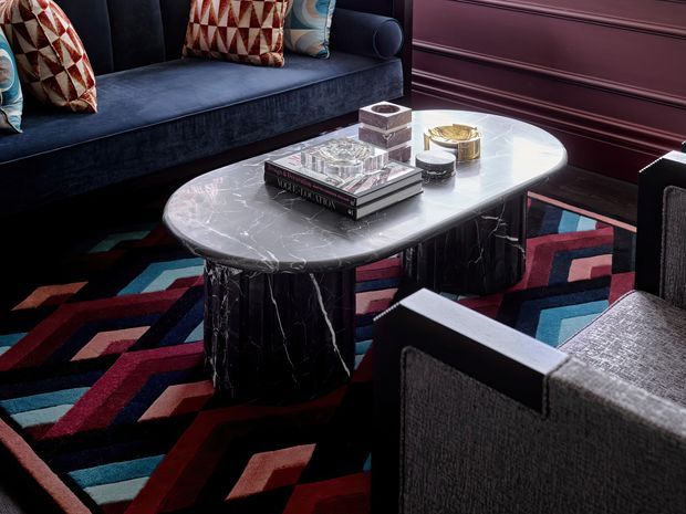 A sophisticated, moody lounge seating area with a deep navy upholstered sofa and matching armchairs arranged around a low oval marble coffee table. The room features dark burgundy wall panelling, a bold geometric multicoloured rug, patterned cushions, and carefully styled coffee-table accessories. The overall composition is richly layered and strongly Art Deco in character.