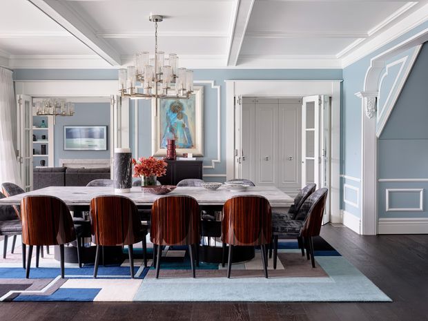 A refined formal dining room with a long marble dining table surrounded by dark, sculptural upholstered chairs. The room features pale blue wall panelling with crisp white moulded trim, dark timber flooring, a coffered ceiling, an ornate multi-arm glass chandelier, and classical decorative detailing. A living area is visible through the open connection at the rear.