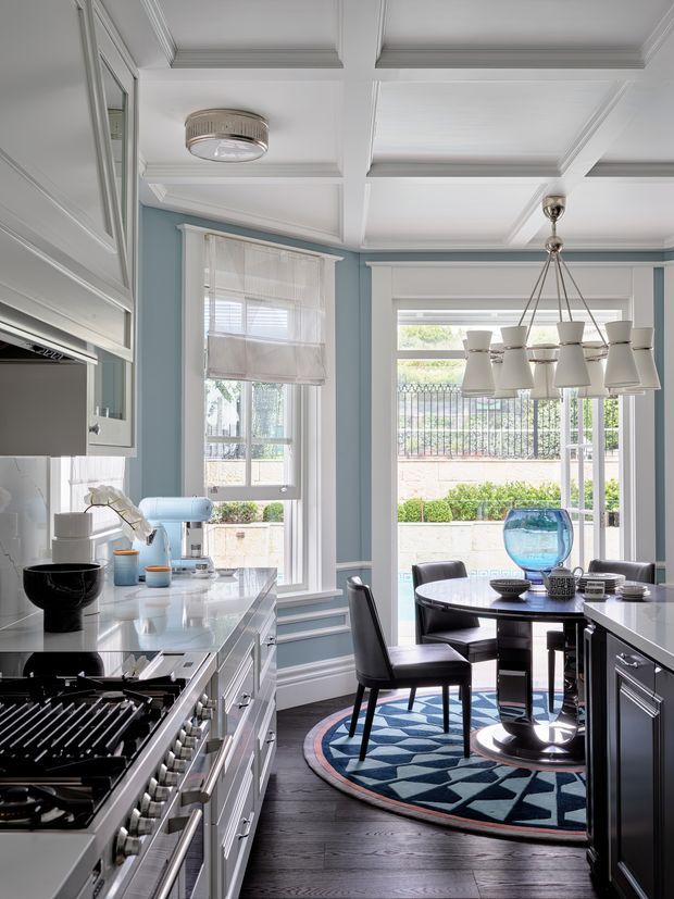 A refined kitchen and dining area with pale blue walls, white cabinetry, dark timber flooring, and a detailed coffered ceiling. The kitchen includes a large gas range, pale stone-look benchtops, and integrated cabinetry. A round dining table with black chairs sits beside tall windows and glazed doors, beneath a multi-light pendant chandelier. The room combines crisp traditional detailing with contemporary furnishings and strong natural light.