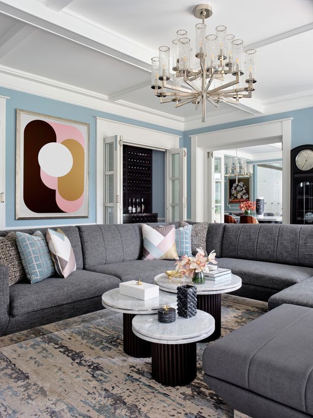 Elegant living room with a large charcoal-grey sectional sofa arranged around layered round marble-top coffee tables. The room features pale blue walls, white decorative ceiling moulding and trim, a dramatic multi-arm glass-and-metal chandelier, a built-in wine display cabinet, abstract artwork, and a patterned area rug. A bright adjoining room is visible through a wide opening.