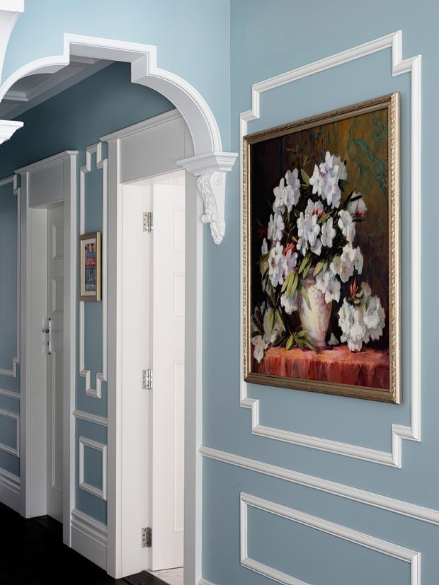 Elegant traditional hallway with muted blue walls, extensive white decorative wainscoting and panel mouldings, a curved arched doorway with ornate corbels, white paneled doors, and a framed floral painting. The dark floor and strong white trim create a refined, formal interior.