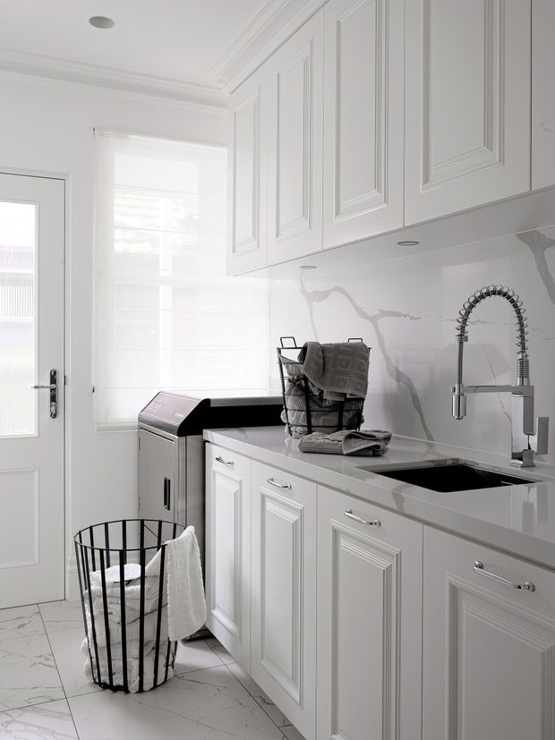 Bright, compact laundry room with white raised-panel cabinetry, a long pale stone benchtop, undermount sink and spring-coil tap, marble-look splashback, and white marble-look floor tiles. A front-loading washing machine, drying rack, and laundry basket reinforce the room's function. The space has classic detailing and a clean monochromatic finish.