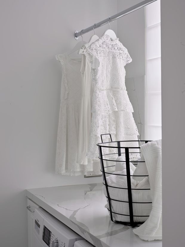 A bright, predominantly white laundry room with a white front-loading washing machine beneath a white marble-look countertop. A black wire laundry basket and folded white textiles sit on the counter, while white garments hang from a wall-mounted rail in front of a tall mirror or reflective panel. The space has a clean, minimal, monochromatic appearance with a small window providing daylight.