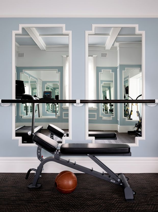 A compact, refined home gym with a black adjustable weight bench and basketball on a dark patterned floor. Pale blue-grey walls feature white decorative moulding and large ornate-framed mirrors, creating repeated reflections and adding depth. A horizontal ballet or exercise barre spans the mirrors, while additional gym equipment is visible in reflection. The room has a bright, classic interior with strong symmetry and polished detailing.