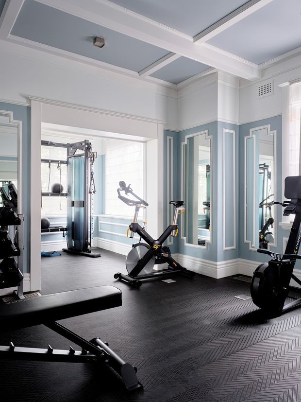 A refined home gym with multiple exercise machines, including a stationary bike, rowing machine, cable machine and treadmill-like equipment. The room features a dark patterned rubber floor, pale blue walls, extensive decorative white wall mouldings and panels, full-height mirrors, a coffered ceiling, and bright windows.