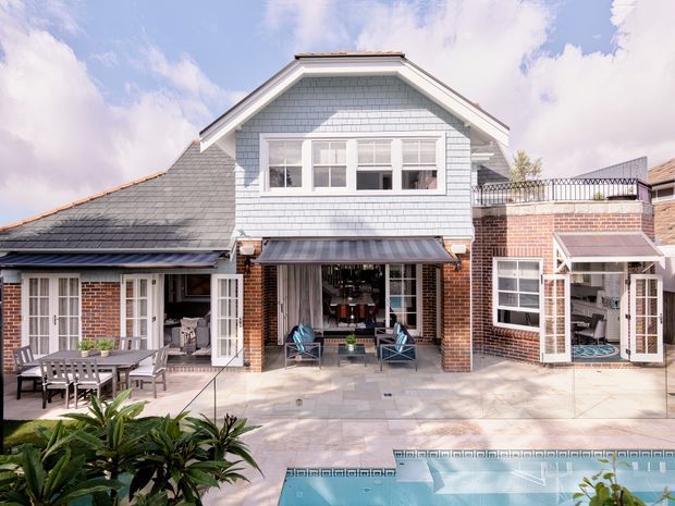 A bright rear exterior view of a multi-level suburban residence with a central gabled section, pale shingle cladding, red brick walls, dark tiled roofing, and multiple glazed doors opening onto a paved courtyard. Outdoor dining and lounge furniture sit beside a rectangular swimming pool, with a rooftop terrace and metal balustrade visible on the right.