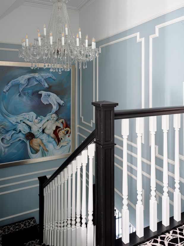Elegant residential stair hall with a traditional painted balustrade and dark-stained timber handrail and newel posts. Pale blue walls are articulated with extensive white decorative moulding and panel trim. A large ornate crystal chandelier hangs above the stair, while a dramatic blue-toned figurative painting forms a focal point on the wall. A patterned black-and-white stair runner is visible at the lower right.