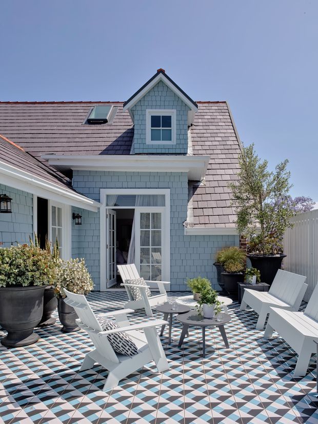 A bright courtyard terrace beside a blue shingle-clad building, with a steep gabled dormer, tiled roof, white-trimmed glazed doors, patterned geometric paving, and white outdoor lounge chairs around small tables and planters.