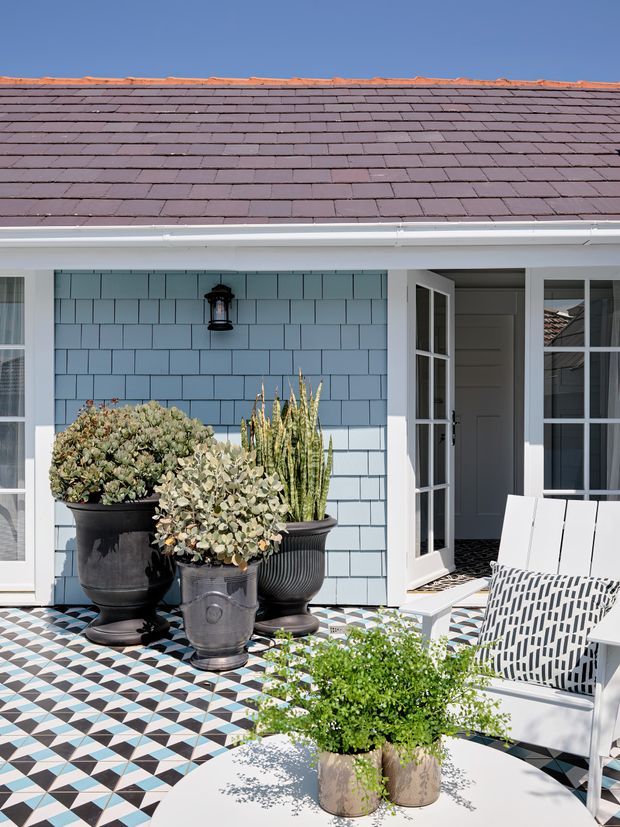 Bright exterior view of a coastal-style home patio or entry terrace. The facade has pale blue shingle cladding, white trim, a dark slate-tile roof, French doors, and a striking geometric black, white, and blue tiled floor. Large potted succulents and white outdoor lounge chairs create a refined, colourful outdoor setting.