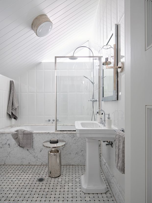 Bright, compact white bathroom with a sloped, panelled ceiling, tiled walls, freestanding pedestal basin, glass-enclosed shower, and a built-in bathtub clad in veined marble. Small monochrome mosaic floor tiles, chrome fixtures, wall mirror, towels, and a small metallic side table create a refined traditional interior.