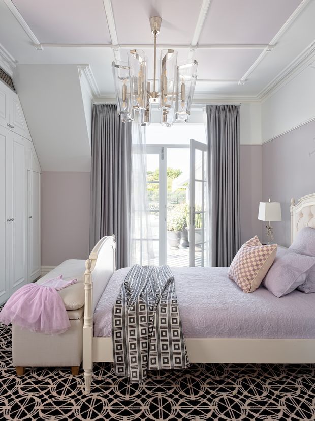 Bright, refined bedroom with pale lavender walls and bedding, white traditional panelled cabinetry, an ornate white bed frame, full-height grey curtains, French doors opening to a balcony or terrace, and a large geometric black-and-cream carpet. A sculptural crystal pendant light forms the focal point overhead, with soft feminine furnishings and layered textiles throughout.