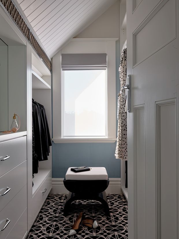 A compact, refined walk-in wardrobe or dressing room with built-in white cabinetry, open hanging storage, a central upholstered ottoman, patterned dark rug, and a tall window. The room features a steeply sloped white-panelled ceiling, pale blue-grey lower wall panelling, white trim, and a detailed panelled door. Natural light creates a calm, tailored atmosphere.