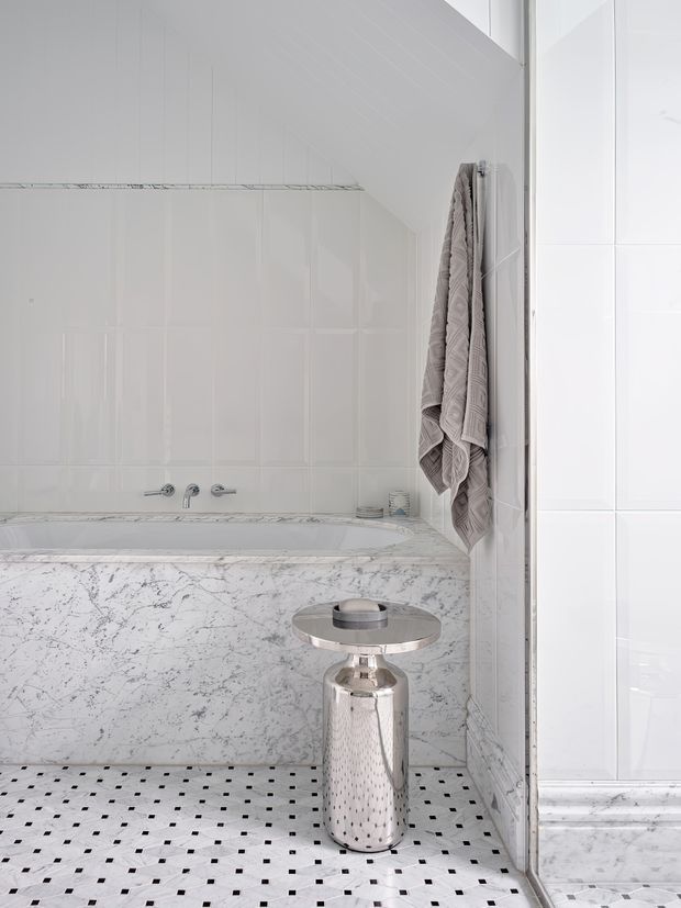 Bright, refined bathroom featuring a built-in marble-clad bathtub surround, white vertically panelled and tiled walls, and a white mosaic tile floor with small black square accents. A large mirror reflects the tub area, while a chrome pedestal side table and neatly hung textured towels add metallic and soft grey details.