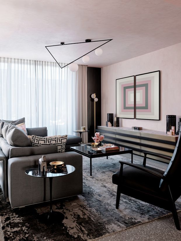 A sophisticated contemporary living room with a charcoal sectional sofa, black armchair, glass coffee tables, a long low media console, geometric framed artwork, and a textured area rug. Full-height sheer curtains provide soft daylight, while a sculptural linear ceiling light and dark vertical wall panelling add architectural interest. The palette is predominantly grey, black, blush pink, and muted metallic tones.