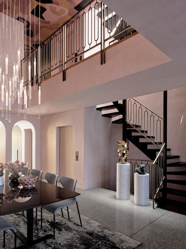 A sophisticated double-height interior featuring a dining area and sculptural black staircase. Decorative metal balustrades with repeated rounded-rectangle motifs line the upper landing and stair, while long suspended glass pendant lights descend beside the dining table. The space uses pale plaster walls, a polished dark dining table, terrazzo-style flooring, marble pedestal columns, and contemporary sculptural decor in a refined, dramatic palette.