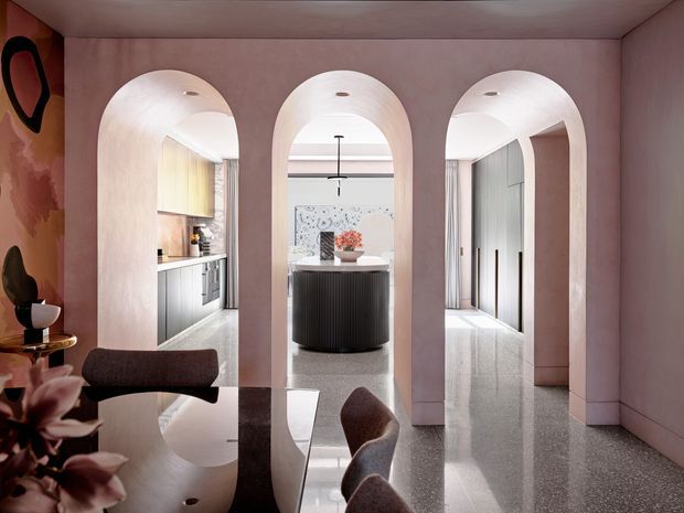 Refined contemporary interior with a blush-pink plastered hallway and three rounded arch openings leading toward a kitchen and central island. A glossy dark dining table and upholstered chairs occupy the foreground, while terrazzo-style flooring, soft curtains, sculptural lighting, and dark fluted cabinetry add contrast.