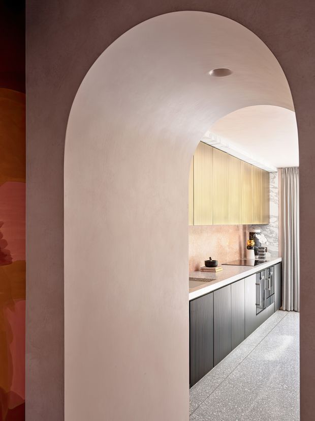 A refined contemporary kitchen viewed through a large rounded archway. The kitchen features vertically ribbed dark cabinetry, pale wood upper cabinets, a light stone benchtop and splashback, terrazzo-style flooring, marble-patterned wall detailing, and full-height curtains. The surrounding arch and walls have a softly textured blush-pink plaster finish, creating a warm, sculptural transition into the kitchen.