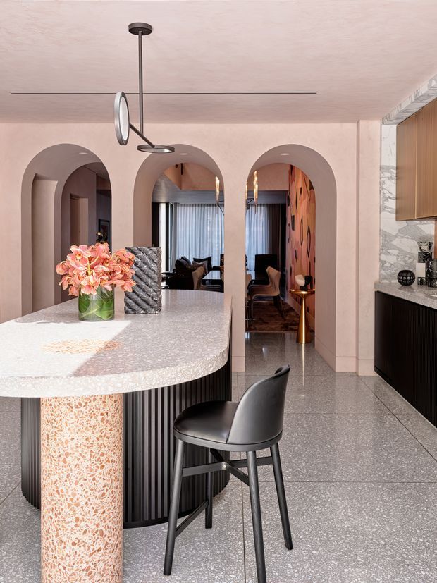 A refined contemporary kitchen with a curved terrazzo island and matching cylindrical support, dark fluted cabinetry, and a pale stone or terrazzo benchtop. Soft pink plaster walls feature three arched openings leading to adjoining living and dining areas. The kitchen includes marble-patterned splashback or wall cladding, timber cabinetry, polished grey terrazzo flooring, sculptural pendant lighting, and a black bar stool. Warm, luxurious finishes create a cohesive statement interior.