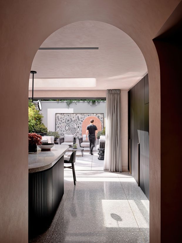 A contemporary open-plan interior viewed through a softly curved arched opening. The foreground contains a dark ribbed kitchen island with a speckled stone or terrazzo benchtop and bar seating. Beyond is a bright living area with pale upholstered seating, indoor plants, artwork, full-height curtains, and dark built-in cabinetry. The space has a softly plastered curved ceiling and walls, large tiled flooring, and strong natural light creating a calm, refined atmosphere.