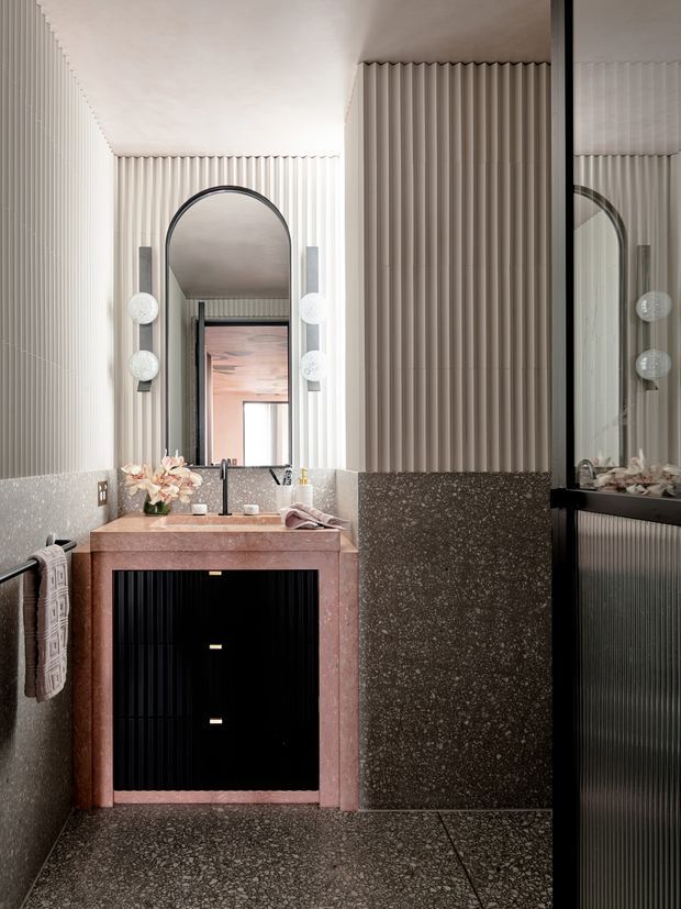 A refined contemporary bathroom featuring a compact pink-toned stone vanity with black fluted cabinetry, a round arched mirror, globe wall lights, terrazzo-style dark flooring and lower wall surfaces, and vertically fluted light wall panelling. A glass shower enclosure is visible at right through reflection.