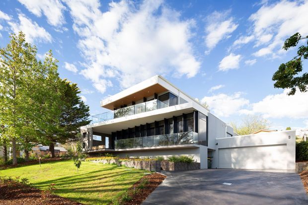 Contemporary multi-storey residence with crisp white geometric forms, broad projecting balconies, extensive glass balustrades, dark-framed floor-to-ceiling glazing, and a large integrated double garage. The home sits on a landscaped suburban site with a gently sloping driveway and mature trees under a bright blue sky.