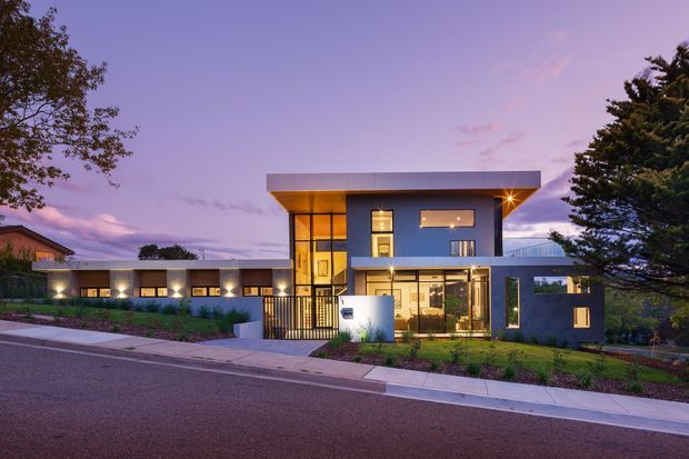 Twilight exterior view of a contemporary multi-level residence with a strongly rectilinear composition, broad flat roof overhangs, dark rendered walls, extensive glazing, and a glazed entry volume. The home sits behind a landscaped front garden with a pedestrian gate and paved street frontage; warm interior lighting contrasts with the cool dusk sky.