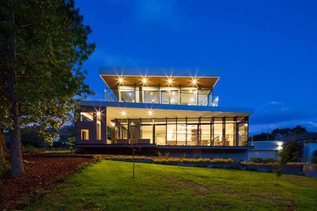 Dusk exterior photograph of a large contemporary two-storey residence with a strongly horizontal composition, flat projecting roof planes, extensive floor-to-ceiling glazing, glass balcony balustrades, and warm interior and exterior lighting. The home sits within a landscaped lawn and garden, with mature trees framing the left side.
