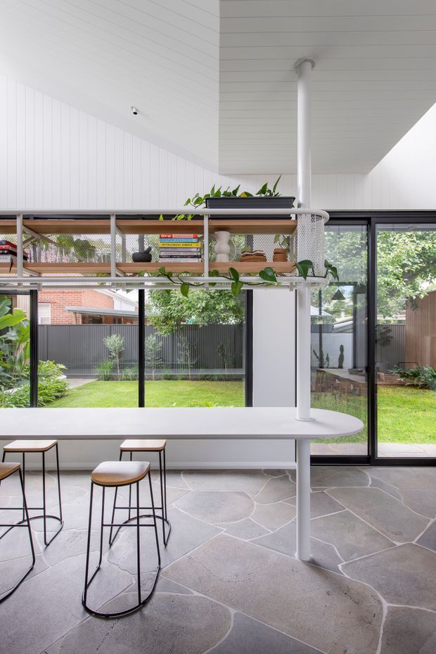 Bright contemporary kitchen or living-kitchen space with a long white peninsula counter, slim black-framed stools, open timber shelving with books and trailing plants, and extensive floor-to-ceiling glazing overlooking a landscaped courtyard. The ceiling and upper walls are clad in white vertical boards, while the floor has large irregular natural stone slabs. A prominent white cylindrical structural post supports the ceiling and counter.