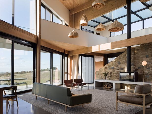 A spacious, light-filled contemporary living room with a double-height ceiling and mezzanine-like upper opening. Floor-to-ceiling glazing frames a rural landscape, while exposed timber ceiling beams and trim contrast with dark metal window frames and a substantial rough-stone fireplace wall. The room includes a modern sofa, leather lounge chairs, a textured area rug, sculptural woven pendant lights, and built-in firewood storage. A glazed roof section brings additional daylight into the open-plan space.