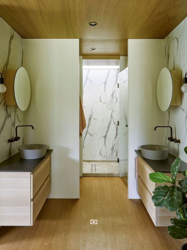 A symmetrical contemporary bathroom with twin floating timber-veneer vanities, vessel basins, matte black wall-mounted taps, oval mirrors and globe wall lights. Pale marble-look wall surfaces frame a central walk-in shower, while the warm timber ceiling and flooring create a cohesive, refined fit-out. White fluted wall panels and indoor greenery add texture and softness.