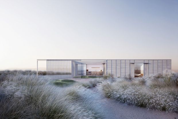 A low, elongated modern residence set within dense coastal dune grasses. The building has a highly rectilinear composition with a flat roof, extensive floor-to-ceiling glazing, pale horizontal and vertical panel cladding, and a recessed central entry or living zone. A sandy path leads through the landscaped grasses toward the house.