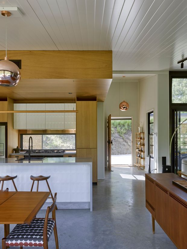 A bright open-plan kitchen and dining area with warm timber cabinetry and ceiling panels, a white island with integrated cooktop and sink, and a timber dining table with sculptural stools and patterned seating. The space has a polished concrete floor, large dark-framed windows and doors, pendant lights, and a view through the open entry to the landscaped exterior. The interior combines clean modern lines with warm mid-century-inspired furniture and detailing.