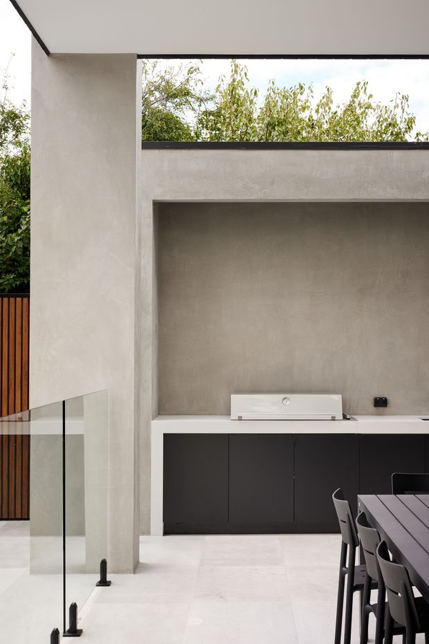 Contemporary outdoor entertaining area with a rendered concrete or stucco wall, long built-in dark cabinetry and benchtop, a white built-in barbecue, and a large dining table with dark chairs. A frameless glass balustrade borders the space, while a narrow clerestory-like opening brings in views of greenery. The restrained palette of warm grey, white, black, and timber creates a refined, minimalist feel.