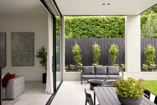 A contemporary indoor-outdoor living area with large black-framed sliding glass doors opening from a bright interior lounge onto a covered patio. The patio includes a grey outdoor sofa, round coffee tables, a dark dining table with chairs, built-in planter beds, and a manicured garden enclosed by a dark vertical timber fence. The interior has pale tiled flooring, a light neutral wall palette, framed artwork, and a grey sofa with a red cushion.