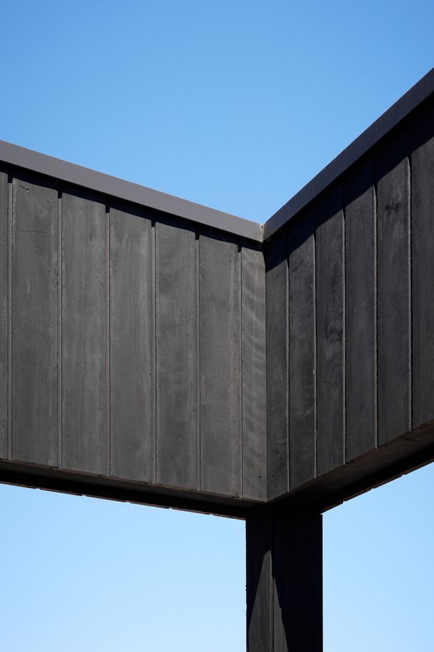 Close-up exterior architectural detail showing a dark, sharply projecting corner or cantilevered box form. The facade is clad in vertically divided charcoal-grey panels with dark trim and a substantial supporting post beneath, silhouetted against a clear blue sky. The image emphasizes geometric massing, exposed structure, and a high-contrast modern exterior.
