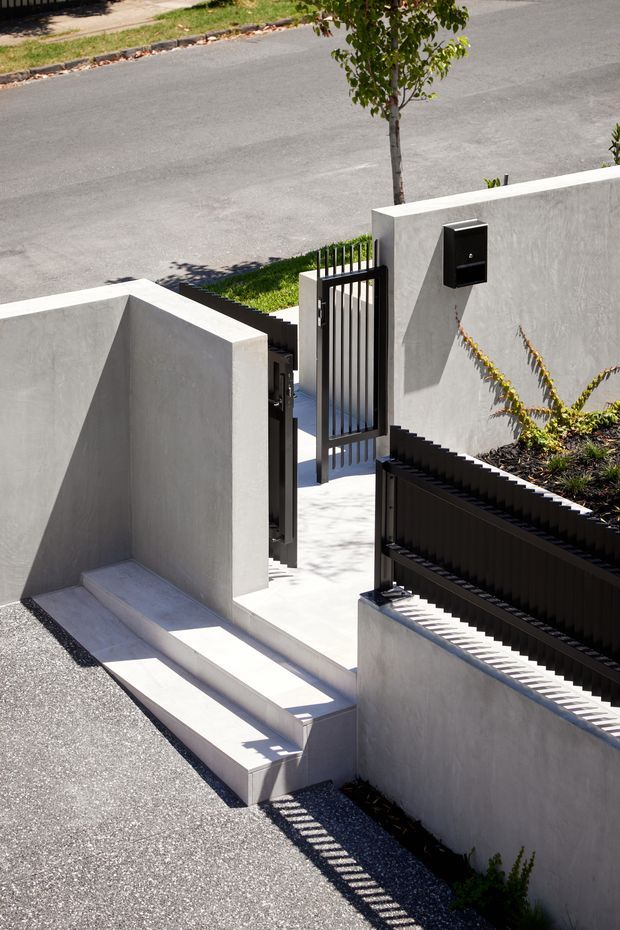 Sunlit exterior entry approach with pale rendered concrete walls, a short flight of concrete steps, a narrow paved pedestrian path, and a black vertical-slat metal gate and fence. A black wall-mounted letterbox is visible beside the gate, with a quiet residential street beyond.