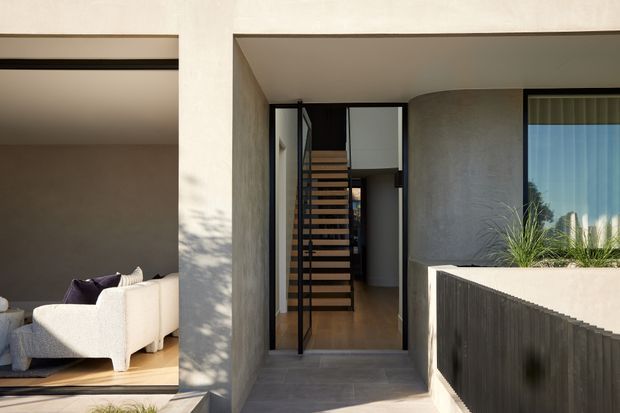 Contemporary residential entrance viewed from outside, featuring a recessed covered entry, dark-framed glazed door, timber floating staircase visible inside, rendered concrete-toned walls, and a raised planter with ornamental grasses. A living room with a light sofa is visible through the adjacent opening.