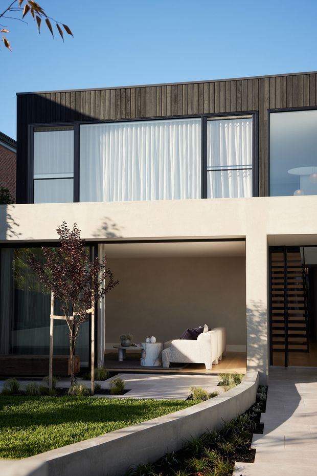 Contemporary two-storey residential facade with dark vertical timber cladding, expansive black-framed glazing, and a pale rendered concrete balcony band. The ground level opens onto a landscaped courtyard with a sheltered outdoor living area furnished with a light sofa and coffee table. A curved raised planter and concrete pathway lead toward the entry, while a horizontal timber slat screen defines the doorway.