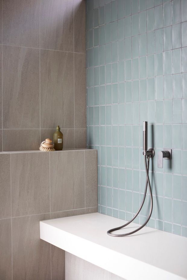 Close-up of a contemporary bathroom shower area, featuring pale blue-green square wall tiles, large warm grey stone-look tiles, a white built-in bath or shower ledge, and a chrome handheld shower fitting. A recessed shelf holds a soap dispenser and brush.