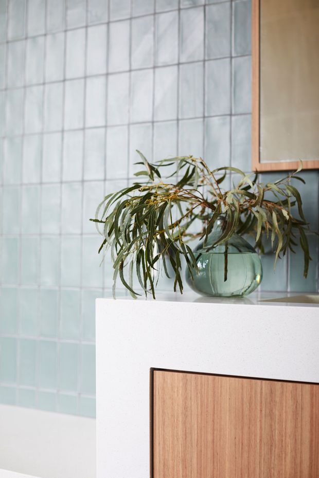 Close-up of a refined bathroom vanity with a pale speckled stone benchtop, warm timber drawer or cabinet front, and a rounded glass vessel holding leafy branches. The backdrop is a grid of soft pale blue-green square wall tiles, with a partial timber-framed mirror visible at upper right. The composition is softly lit and calm, combining natural materials with a contemporary colour palette.