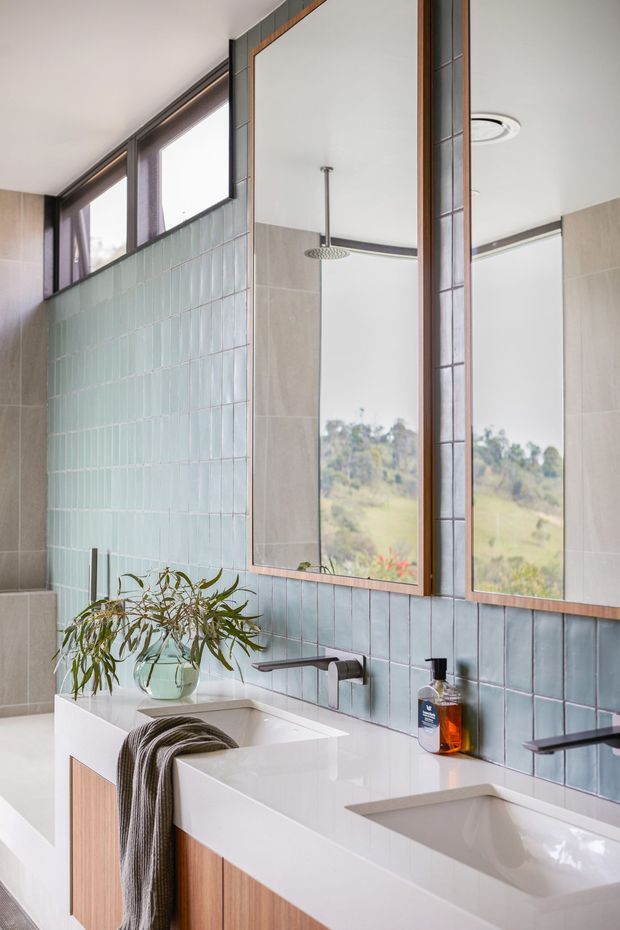 Bright contemporary bathroom with a long double vanity, white solid-surface benchtop, integrated basins, timber-finished cabinetry, and wall-mounted dark metal taps. Pale blue vertical wall tiles form a feature backsplash, with large framed mirrors, a glass shower enclosure, and a freestanding bathtub visible at left. Natural light enters through high windows, creating an airy, refined interior.