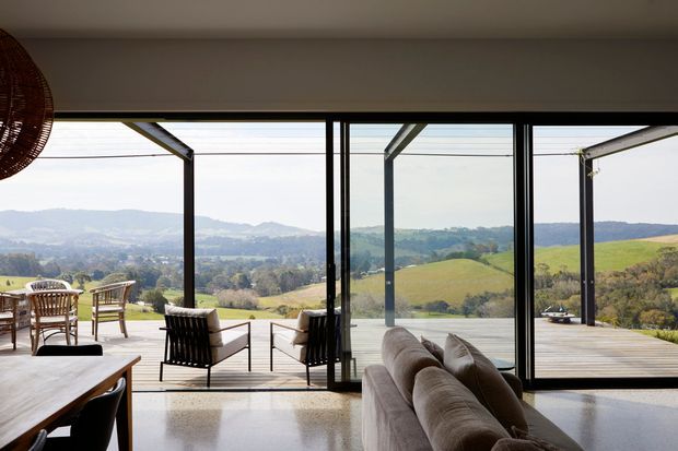 A contemporary open-plan living area with floor-to-ceiling black-framed glazing opening onto a timber deck. The room contains a large neutral sectional sofa, timber coffee table, and woven outdoor-style seating, with expansive views across rolling green countryside and hills. The glazing and open doors create a strong indoor–outdoor connection.