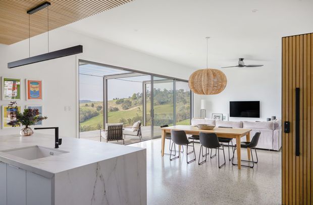 Bright open-plan interior combining a white kitchen with a large waterfall island, dining area, and living room. Full-height glazed sliding doors open to a rural landscape, while timber slatted ceiling detail, a timber slatted entry screen, terrazzo-like flooring, and a woven pendant add warmth to the contemporary space.