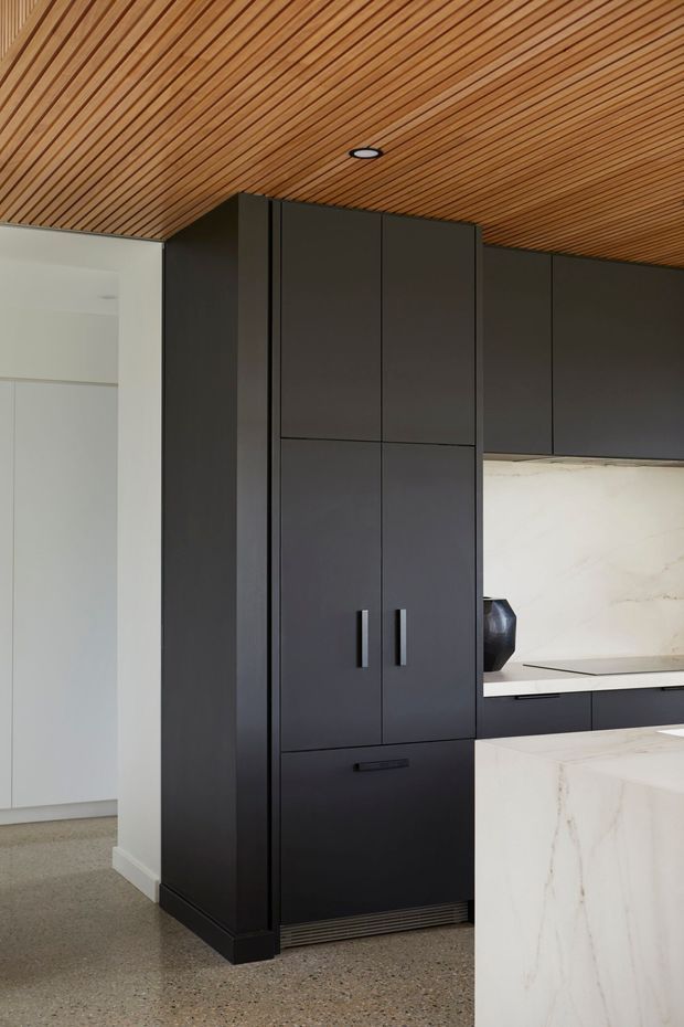 A refined modern kitchen featuring tall matte-black cabinetry, integrated appliances, a pale marble-look splashback and island, and a warm linear timber-slat ceiling. The space has a minimalist, high-contrast palette with polished concrete or terrazzo-style flooring and clean, handle-minimal joinery.