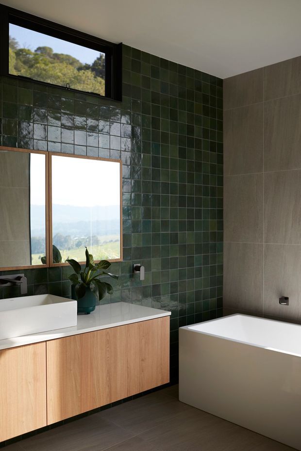 A contemporary bathroom with a floating light-wood vanity, white vessel basin, and freestanding white bathtub. The walls combine glossy dark green square tiles with large warm grey-beige tiles, while timber-framed glazing overlooks a leafy landscape. A potted plant adds a natural accent.