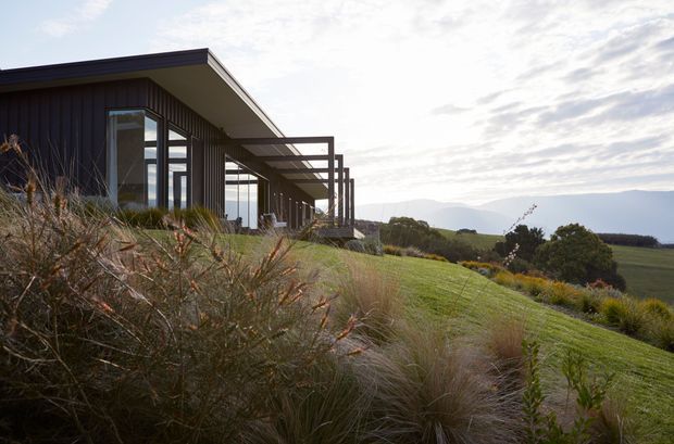 A low, modern single-storey house set into a gently sloping rural landscape. The dark vertically clad facade has expansive glazing and a projecting pergola-like timber frame, opening toward landscaped grasses and distant rolling hills. The restrained architecture contrasts with the natural planting and open countryside.