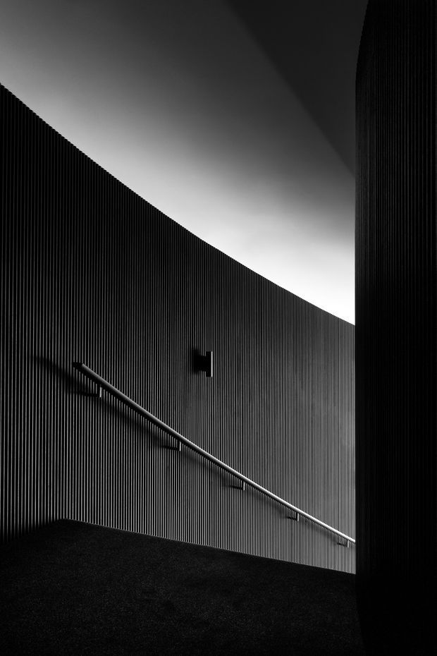 A dramatic monochrome architectural detail showing a narrow ramp or stairway enclosed by vertically ribbed dark walls. A simple metal handrail follows the incline, while a sharply angled opening above admits soft light and creates strong geometric shadows. The image emphasizes texture, contrast, and minimal contemporary form.